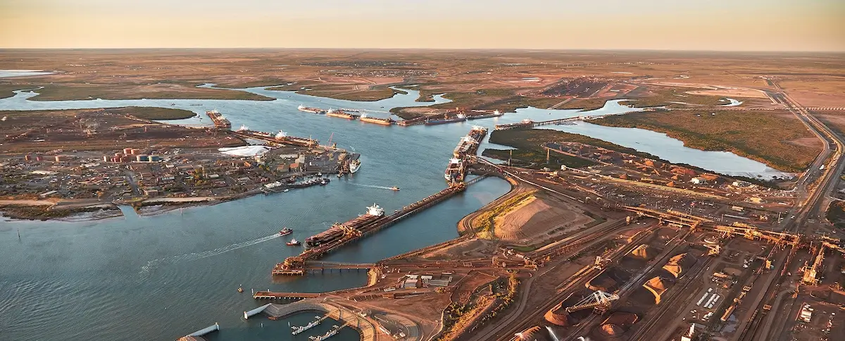 Aerial view of a large industrial port with docks, ships, cranes, and warehouses along a winding river, surrounded by expansive flatlands and scattered infrastructure under a clear sky.