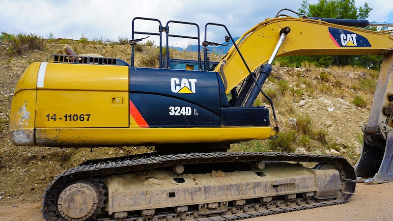 A yellow Caterpillar 324D L excavator with black tracks is parked on a dirt construction site, with grass and a hill in the background.