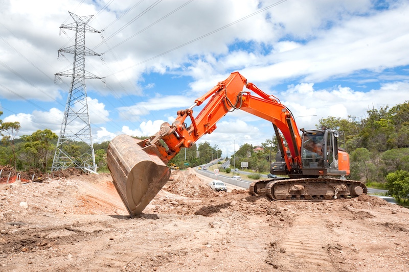 An orange excavator moves dirt on a construction site near a road, with power lines and greenery in the background under a partly cloudy sky.