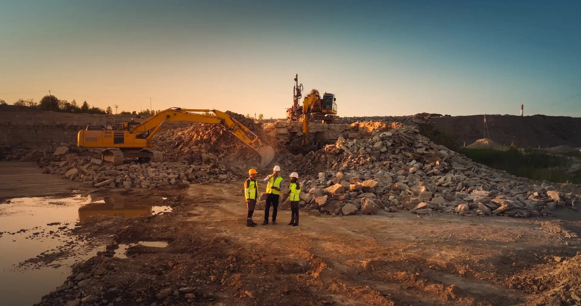 Three construction workers in high-visibility vests and helmets stand on rocky ground, talking near heavy machinery and large piles of rubble at a construction site during sunset.