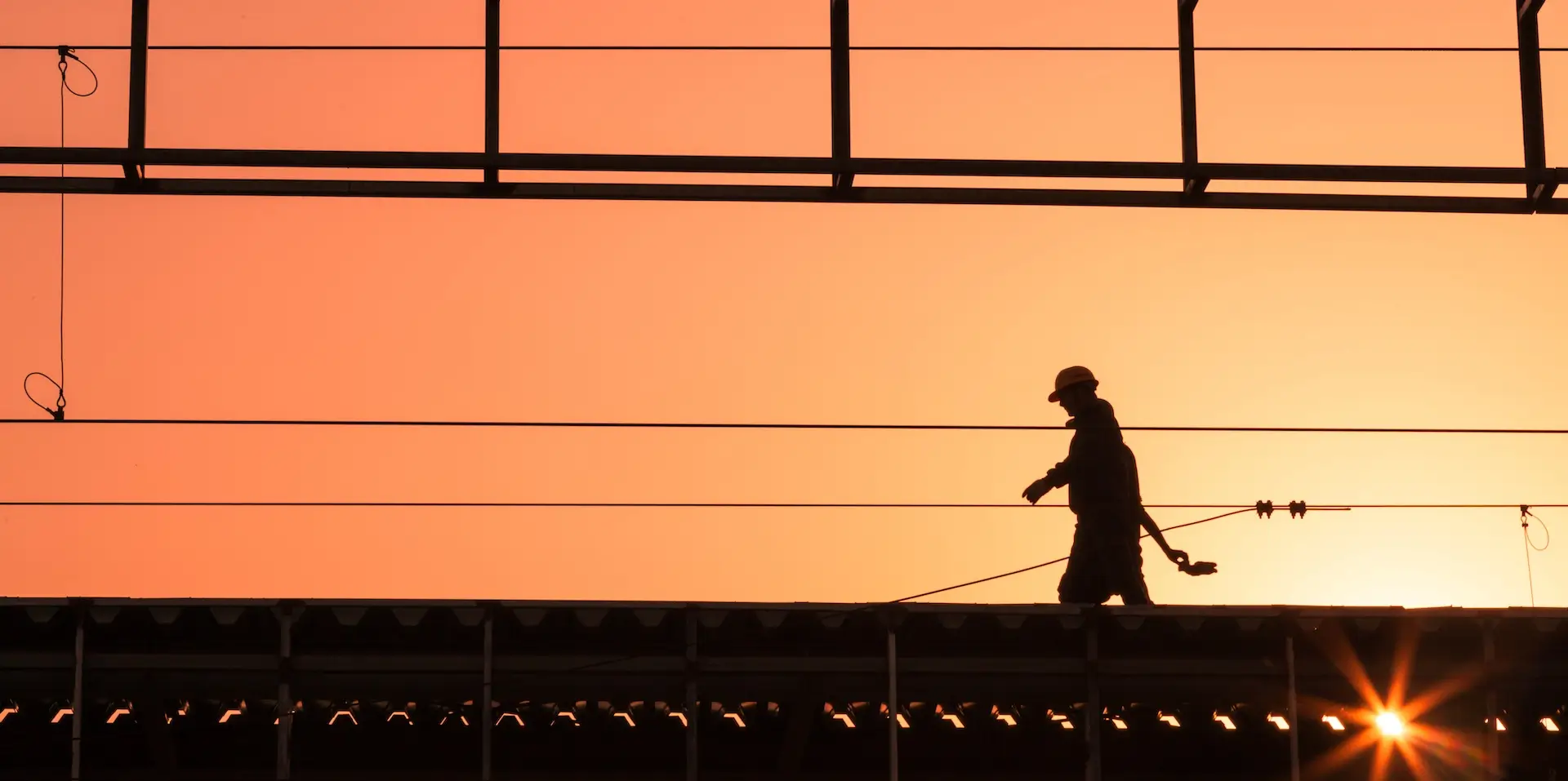 Silhouette of a construction worker walking on scaffolding at sunset, with an orange sky in the background and structural beams overhead.