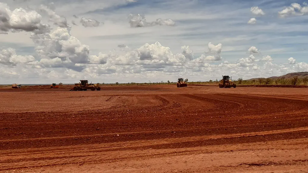 Local civil contractors Pilbara operate construction vehicles on a wide, flat area of reddish-brown earth under a partly cloudy sky, with sparse vegetation and distant hills in the background.
