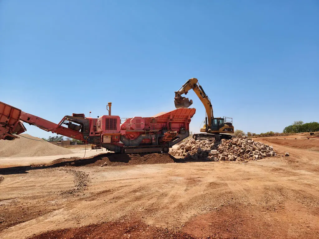 A yellow excavator is loading rocks into a large red rock crusher at a construction or mining site under a clear blue sky, with piles of dirt and gravel—typical of projects handled by local civil contractors Pilbara.