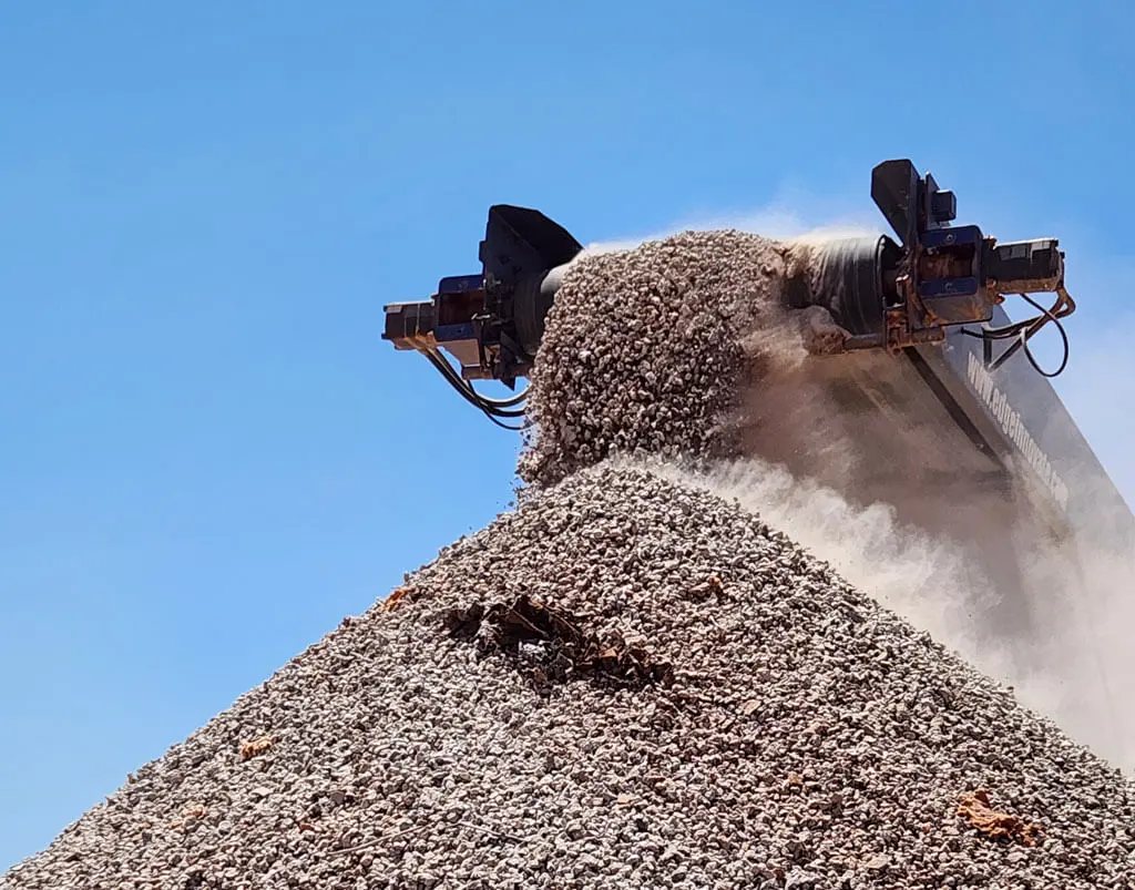 A conveyor belt deposits a large pile of gravel or crushed stone outdoors under a clear blue sky, creating a conical mound as dust rises—routine worksite activity for local civil contractors Pilbara.