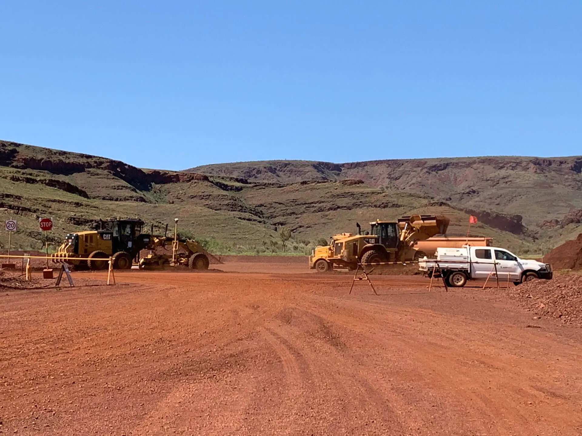 Heavy construction vehicles and a white utility truck are parked on a wide, red dirt road, framed by rocky hills and blue sky—capturing the essence of civil construction jobs Pilbara region is known for. Mining or roadwork activity is suggested.