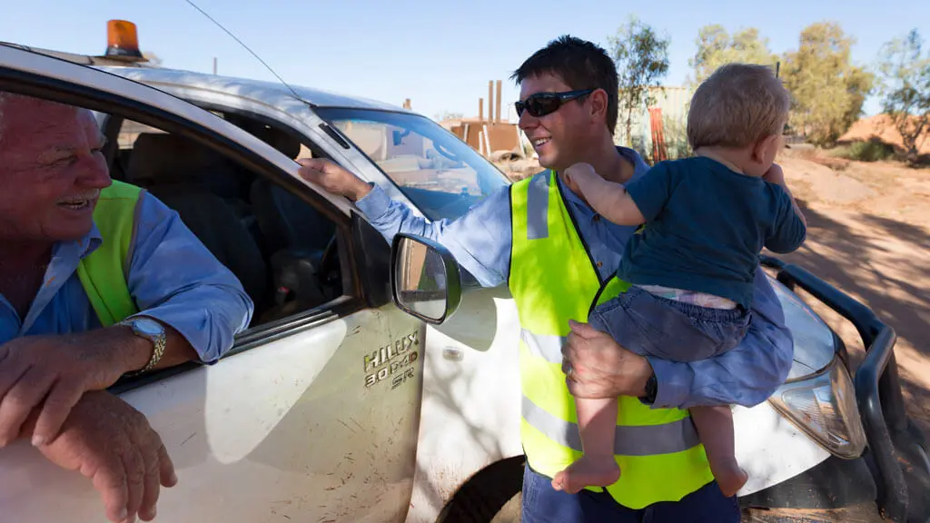 A man in a high-visibility vest, likely one of the local civil contractors Pilbara relies on, holds a young child and smiles while chatting with another man in a white pickup truck on a sunny, dusty rural road.